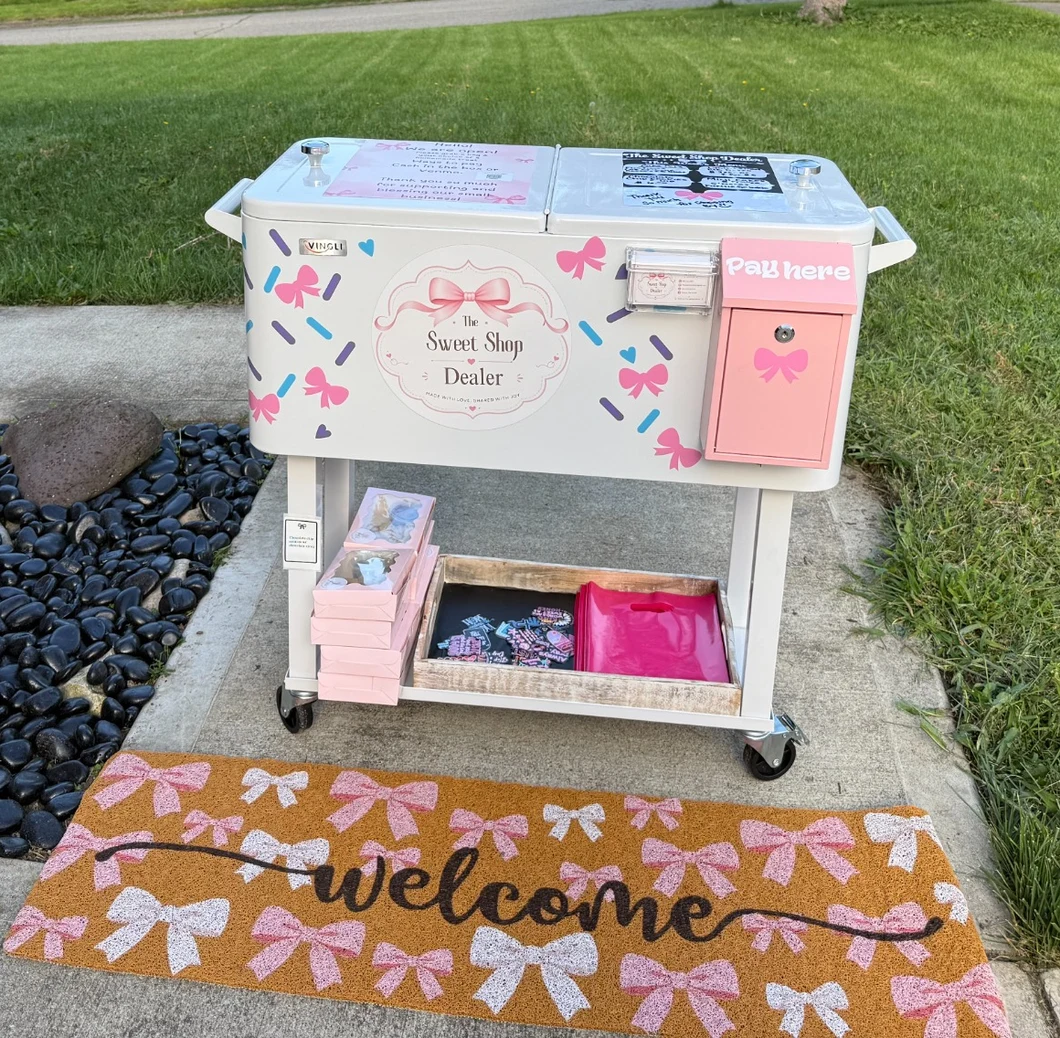The Sweet Shop Dealer’s white outdoor sweet stand, decorated with pink bows, a pink payment box, and a welcome mat.